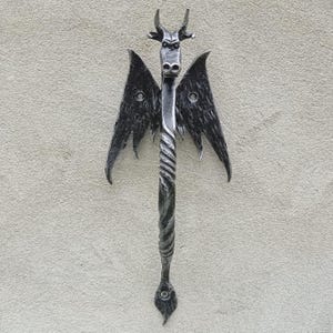 Könnte beinhalten: Handgefertigter Türgriff aus schwarzem Metall. Der Griff hat ein stilisiertes Drachendesign mit Hörnern, Flügeln und einem gedrehten Mittelschaft. Die Flügel haben Nietendetails. Der Griff ist auf einer hellgrauen Oberfläche montiert.