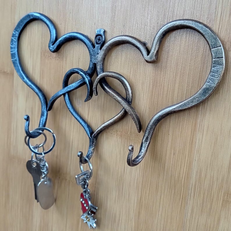 May include: A wall-mounted key holder with three intertwined heart-shaped hooks. The metal key holder has a rustic finish with a mix of silver and bronze tones. Two sets of keys hang from the hooks.