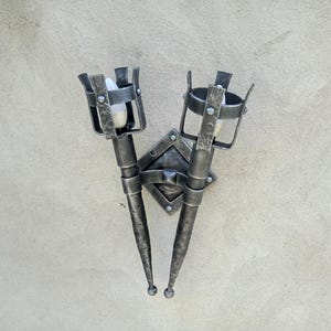 May include: A dark grey wrought iron wall sconce featuring two torch-style light holders. The sconce has a diamond-shaped backplate and cylindrical torch holders with decorative metalwork. It is mounted on a light grey wall. The sconce is approximately 30 cm in height.