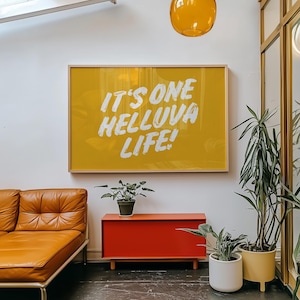 May include: A yellow framed print with the text "It's one helluva life!" hanging on a white wall. A red cabinet with a brown leather couch in front of it. Two potted plants are on the floor.