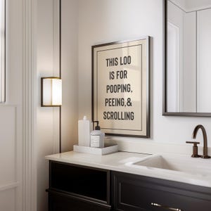 Puede incluir: Un tocador de baño blanco con una encimera negra y un lavabo de porcelana blanca. Una impresión enmarcada con el texto "This loo is for pooping, peeing, & scrolling" cuelga de la pared sobre el tocador. Un espejo enmarcado negro cuelga de la pared sobre el lavabo.