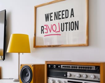 Può includere: Una stampa incorniciata con il testo "We need a revolution" in lettere nere, con la parola "revolution" cancellata e sostituita dalla parola "love" in lettere rosa. Nell'immagine sono presenti anche una lampada gialla, un impianto stereo e uno scaffale in legno.