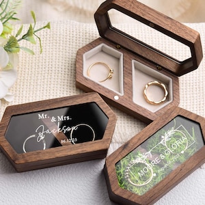 以下が含まれることがあります： 透明な蓋が付いた木製のリングボックスで、結婚指輪を展示しています。1つのボックスは開いており、2つのリングが表示されています。別のボックスには「Mr. & Mrs. Jackson 06.12.25」と書かれています。3番目のボックスには「MOORE & GLORIA August 08, 2024」と書かれています。