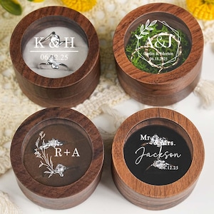 May include: Four wooden ring boxes with clear tops. Two boxes have rings inside, one with "K&H 06.12.25" and the other with "Mr. & Mrs. Jackson 06.12.25". Another box has moss and lights, and the last has "R+A".