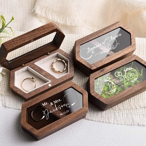 Op de afbeelding: Houten ringendoosjes met doorzichtige deksels, sommige met gegraveerde namen en data. E&eacute;n doosje is open en toont twee gouden ringen. De doosjes zijn bruin met een geometrisch ontwerp. De tekst op de doosjes luidt "Jimmans & Aurora", "Joyce & Ryan" en "Mr. & Mrs. Jackson 06.12.25".
