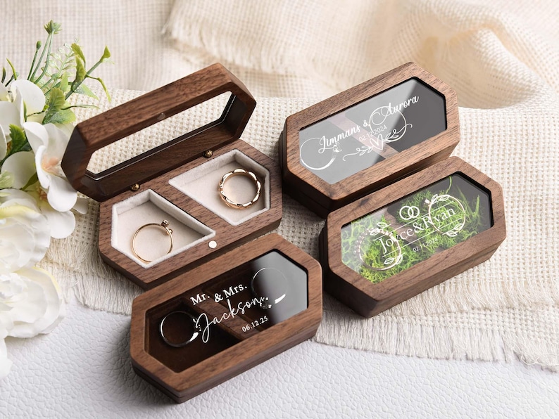Op de afbeelding: Houten ringendoosjes met doorzichtige deksels, sommige met gegraveerde namen en data. E&eacute;n doosje is open en toont twee gouden ringen. De doosjes zijn bruin met een geometrisch ontwerp. De tekst op de doosjes luidt "Jimmans & Aurora", "Joyce & Ryan" en "Mr. & Mrs. Jackson 06.12.25".