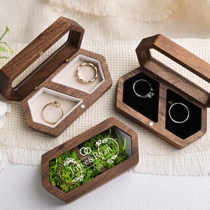 Op de afbeelding: Houten ringendoosjes met doorzichtige deksels, met ringen met gouden en zilveren banden. E&eacute;n doosje heeft de namen "Joyce & Ryan" en een datum. De doosjes zijn open en tonen de ringen.