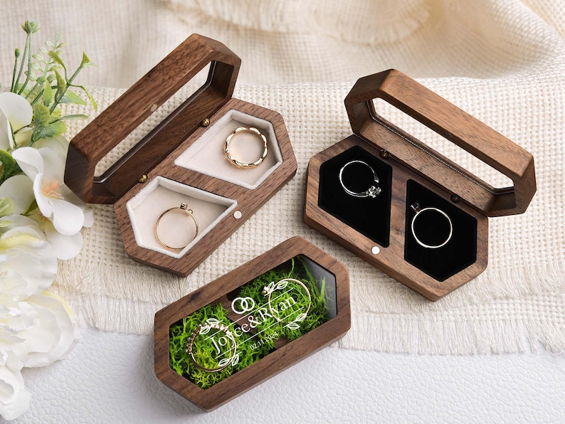 Op de afbeelding: Houten ringendoosjes met doorzichtige deksels, met ringen met gouden en zilveren banden. E&eacute;n doosje heeft de namen "Joyce & Ryan" en een datum. De doosjes zijn open en tonen de ringen.
