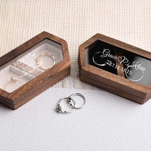Op de afbeelding: Twee houten ringendoosjes met doorzichtige deksels, &eacute;&eacute;n met "Miss & Mrs Smith" en de andere met "Grace & Klein 07.12.2025" gegraveerd. Twee ringen zitten in de doosjes en nog twee eronder. De doosjes staan op een wit oppervlak.