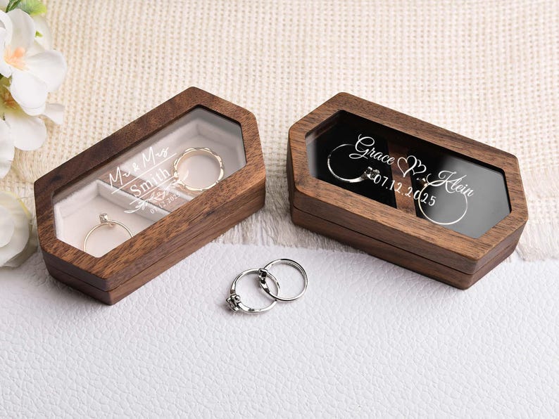Op de afbeelding: Twee houten ringendoosjes met doorzichtige deksels, &eacute;&eacute;n met "Miss & Mrs Smith" en de andere met "Grace & Klein 07.12.2025" gegraveerd. Twee ringen zitten in de doosjes en nog twee eronder. De doosjes staan op een wit oppervlak.