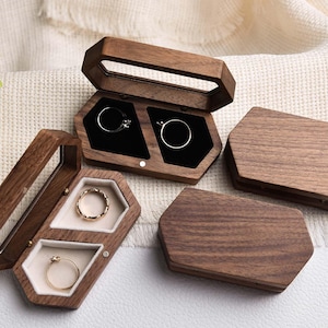 Op de afbeelding: Verschillende zeshoekige houten ringendoosjes, sommige open en andere gesloten. De doosjes zijn gemaakt van donker hout en bevatten ringen met gouden en zilveren banden. De binnenkant is gevoerd met zwarte of cr&egrave;mekleurige stof.