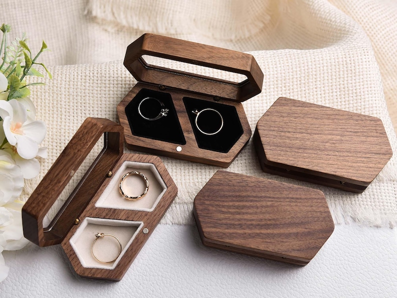 Op de afbeelding: Verschillende zeshoekige houten ringendoosjes, sommige open en andere gesloten. De doosjes zijn gemaakt van donker hout en bevatten ringen met gouden en zilveren banden. De binnenkant is gevoerd met zwarte of cr&egrave;mekleurige stof.