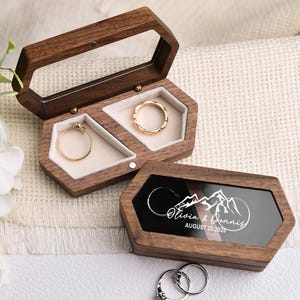 Op de afbeelding: Zeshoekige houten ringendoosjes met doorzichtige deksels, met gouden ringen. E&eacute;n doosje is open en toont twee ringen, terwijl het andere gesloten is en een berglandschap en de namen "Olivia & Dennis" met de datum "AUGUST 20, 2025" toont.