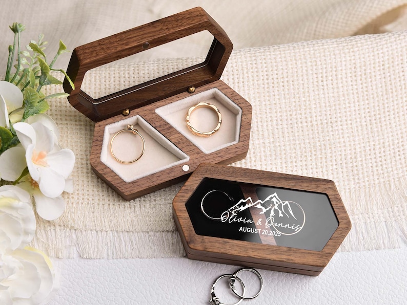 Op de afbeelding: Zeshoekige houten ringendoosjes met doorzichtige deksels, met gouden ringen. E&eacute;n doosje is open en toont twee ringen, terwijl het andere gesloten is en een berglandschap en de namen "Olivia & Dennis" met de datum "AUGUST 20, 2025" toont.