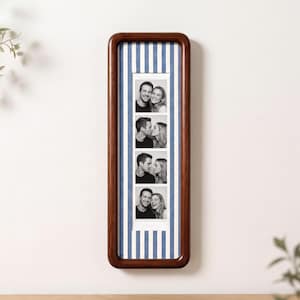 May include: A vertical rectangular dark wood photo frame containing a strip of four black and white photographs of a couple. The background inside the frame features a pattern of vertical blue and white stripes behind the photo strip.