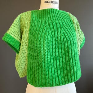 Puede incluir: Un suéter verde y verde claro de manga corta con cuello redondo. El suéter está hecho de un material de punto y tiene un diseño texturizado. La etiqueta en el maniquí dice "SIGURDSDOTTER".