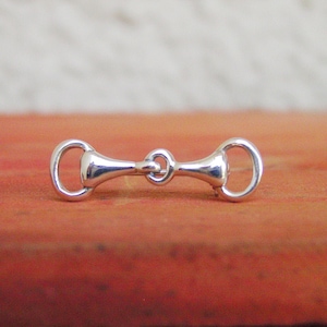 May include: A silver-toned snaffle bit charm, a type of horse bit, with a rounded shape and a smooth finish.