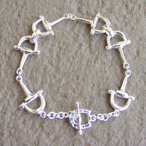 May include: Silver bracelet with a series of linked horse bit charms and a toggle clasp.