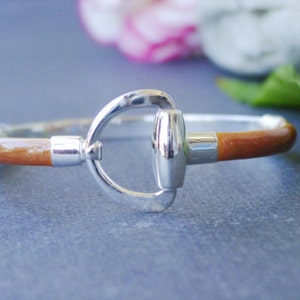 May include: A brown leather bracelet with a silver equestrian-style buckle. The bracelet is a simple design with a smooth leather band and a polished silver buckle.
