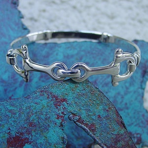 May include: A silver bangle bracelet featuring a snaffle bit design. The bracelet is crafted from polished metal and has a simple, elegant style.