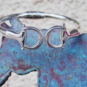 Könnte beinhalten: Ein silbernes Armreif-Armband mit zwei Anhängern in Form von Pferdebits. Das Armband liegt auf einer blau-grünen, strukturierten Oberfläche.