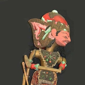 Puede incluir: Un títere tradicional indonesio con una cara pintada de rojo, marrón y dorado y una falda de tela con estampado colorido. El títere sostiene dos palos de madera.