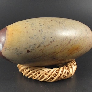 Könnte beinhalten: Ein großer, ovaler Stein in Braun- und Beigetönen mit weißen und roten Markierungen. Der Stein liegt auf einem geflochtenen, runden Ständer.