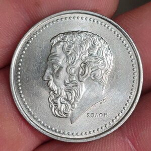 May include: A silver coin featuring a detailed profile of a man with a beard, surrounded by a dotted border. The coin has Greek text and is likely a collectible or historical item.