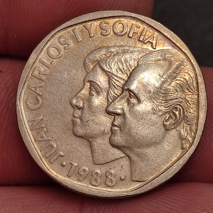 May include: A gold-colored coin featuring profiles of two people, with the text "JUAN CARLOS I Y SOFIA" and the year "1988" around the edge. The coin is held between two fingers.