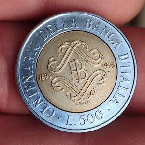 Puede incluir: Primer plano de una moneda italiana de 500 liras. La moneda tiene un anillo exterior plateado y un centro dorado. La moneda presenta el texto "CENTENARIO DELLA BANCA D'ITALIA" y el año 1993. La moneda también tiene el texto "L. 500" y el año 1893.