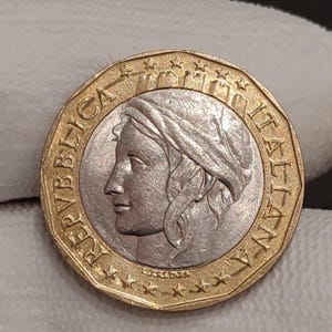 Puede incluir: Una moneda italiana de un euro. Presenta el perfil de una mujer, rodeado de las palabras "REPUBBLICA ITALIANA" y estrellas. El borde exterior es dorado y el centro plateado.