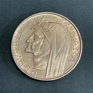 May include: A silver coin featuring a profile of a person wearing a laurel wreath and a draped garment. The coin has the words "REPUBBLICA ITALIANA" and the name "S. VERGINI" inscribed around the edges.