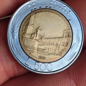 Puede incluir: Primer plano de una moneda italiana de 500 liras de 1985. La moneda presenta una representación detallada de un edificio con una estatua y una persona a caballo. La moneda tiene un anillo exterior plateado y un centro dorado.