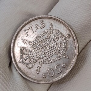 Puede incluir: Una moneda de plata con la inscripción "PTAS" y el número "50". La moneda presenta un emblema detallado en el centro, rodeado por un borde ornamentado. La moneda está sujeta por una mano enguantada de blanco.