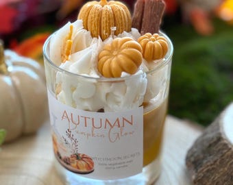 Könnte beinhalten: Eine Kerze im Klarglas mit orangefarbenem Wachs, verziert mit weißem, geschlagenem Wachs, zwei kleinen orangefarbenen Kürbissen und einer braunen Zimtstange. Das Etikett trägt die Aufschrift Autumn Pumpkin Glow, Witchmoon Secrets, 100% vegetable wax, 200 g candle.