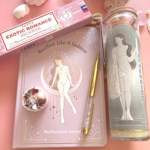 Könnte beinhalten: Eine rosa Zusammenstellung mit einem "Manifest Like A Goddess"-Journal, einem goldenen Stift, einer Kerze mit Aphrodite-Design und einer Schachtel "Exotic Romance"-Räucherstäbchen. Das Journal hat eine nackte Illustration und den Text "Manifestation Journal".