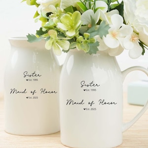 May include: Two white ceramic vases with floral arrangements. One vase is a pitcher with a handle, and the other is a smaller, rounded vase. Both vases have the words "Sister" and "Maid of Honor" printed on them, along with the years "Est. 1995" and "Est. 2025".