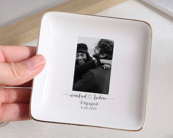 May include: A white ceramic ring dish with a gold rim, featuring a black and white photo of a couple. The dish is inscribed with the names "Winifred & Isidore", the word "Engaged", and the date "6.20.2025".