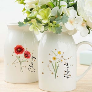 Puede incluir: Dos jarrones de cerámica blanca con diseños florales y nombres. Uno presenta amapolas rojas y el nombre "Cherry", mientras que el otro tiene flores amarillas y el nombre "Roberta". Ambos jarrones contienen flores blancas.