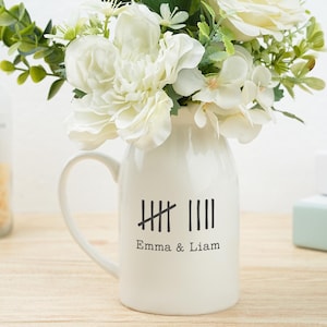 Könnte beinhalten: Eine cremefarbene Keramiktasse mit Henkel, gefüllt mit weißen Blumen und Grünzeug. Die Tasse hat ein Strichlisten-Design und die Namen "Emma & Liam" in Schwarz. Eine Glasflasche und Bücher sind im Hintergrund.