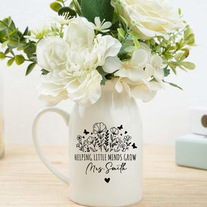 Puede incluir: Un jarrón de cerámica blanco con asa, lleno de flores blancas y vegetación. El jarrón está decorado con un diseño floral negro, mariposas y el texto "Helping Little Minds Grow Mrs. Smith" con un símbolo de corazón.