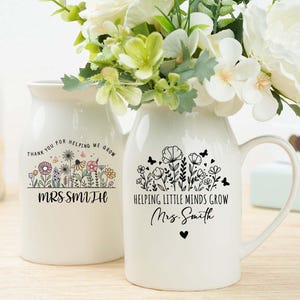 Può includere: Due vasi in ceramica bianca con disegni floreali e testo. Uno dice "Thank you for helping me grow Mrs Smith" e l'altro "Helping little minds grow Mrs Smith". Entrambi i vasi contengono fiori bianchi.