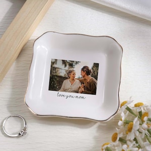 May include: White ceramic trinket dish with a gold rim, featuring a printed photo of two people and the text "love you mom." A silver ring with a small stone sits nearby. Daisies are in the bottom right corner.