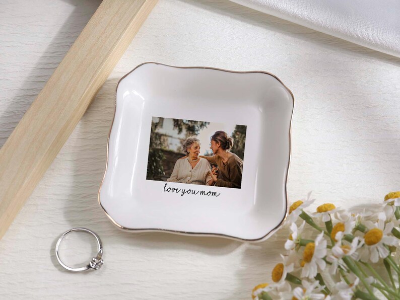 May include: White ceramic trinket dish with a gold rim, featuring a printed photo of two people and the text "love you mom." A silver ring with a small stone sits nearby. Daisies are in the bottom right corner.