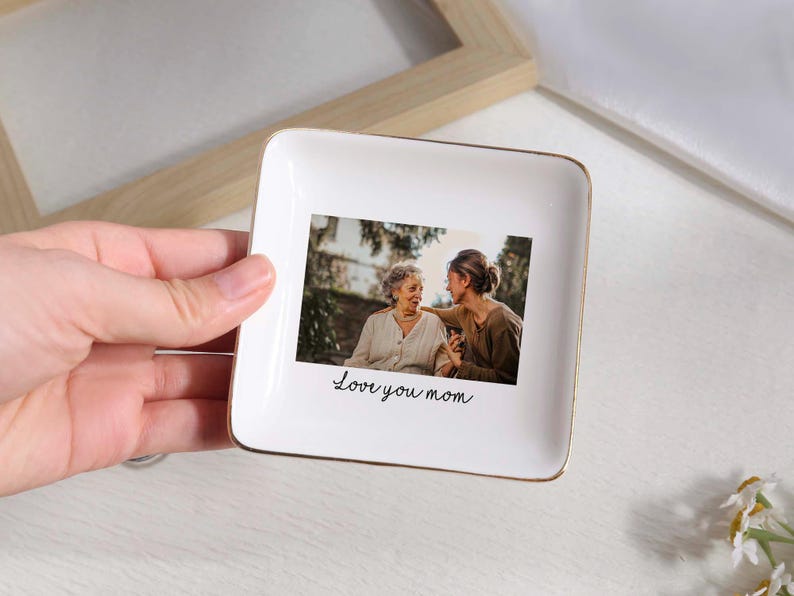 May include: A white square ceramic trinket dish with a gold rim. The dish features a photograph of two women and the handwritten text "Love you mom". The dish is a decorative item.