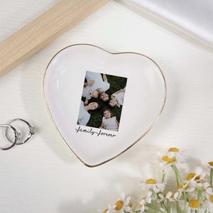 May include: A heart-shaped white ceramic trinket dish with a gold rim, holding a family photo. The photo is surrounded by the words "Family Forever" in script. Two silver rings and a bouquet of daisies are also present.