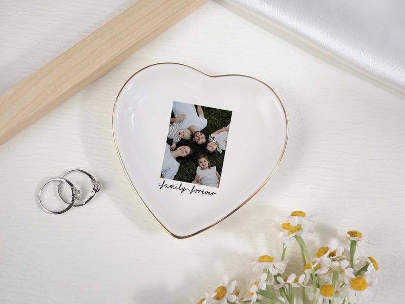May include: A heart-shaped white ceramic trinket dish with a gold rim, holding a family photo. The photo is surrounded by the words "Family Forever" in script. Two silver rings and a bouquet of daisies are also present.