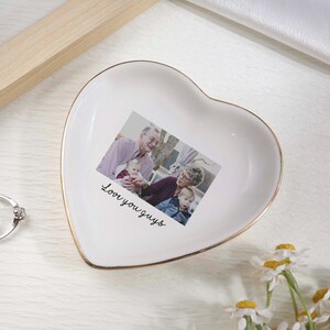 May include: White heart-shaped trinket dish with a gold rim, featuring a family photo and the text "Love you guys." Two silver rings with clear stones are next to the dish. Daisies are in the bottom right corner.