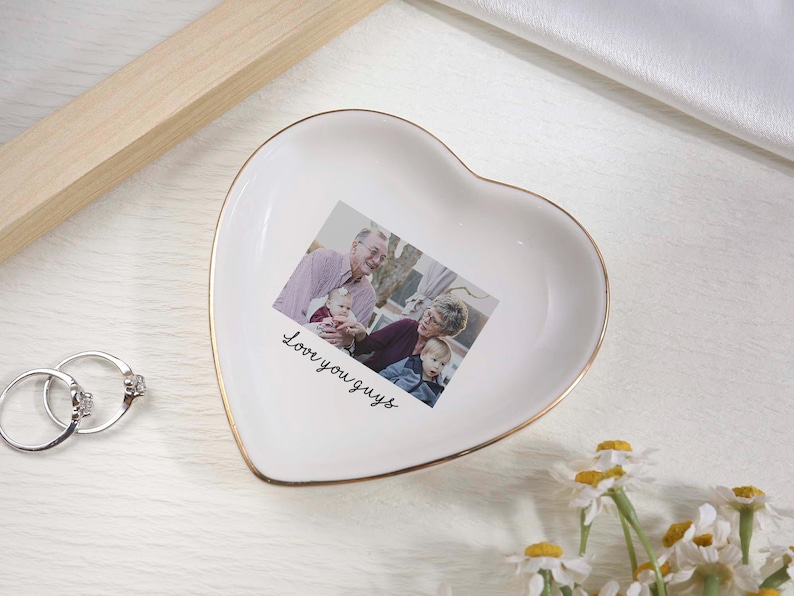 May include: White heart-shaped trinket dish with a gold rim, featuring a family photo and the text "Love you guys." Two silver rings with clear stones are next to the dish. Daisies are in the bottom right corner.