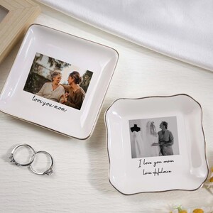May include: Two white ceramic trinket dishes with gold trim, each featuring a photograph and handwritten text. One says "love you mom", the other says "I love you mom Love Heloise". Silver rings are next to the dishes.